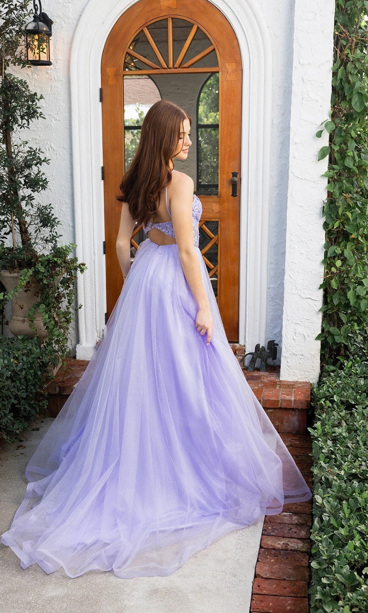 A woman wearing the Portia and Scarlett XO163 prom ball gown with an embellished bodice and glitter tulle skirt stands on a pathway before a wooden arched door, facing away from the camera.